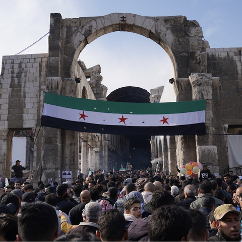 A huge crowd of people gathering outside, in front of a huge Syrian flag hanging from a gate. 