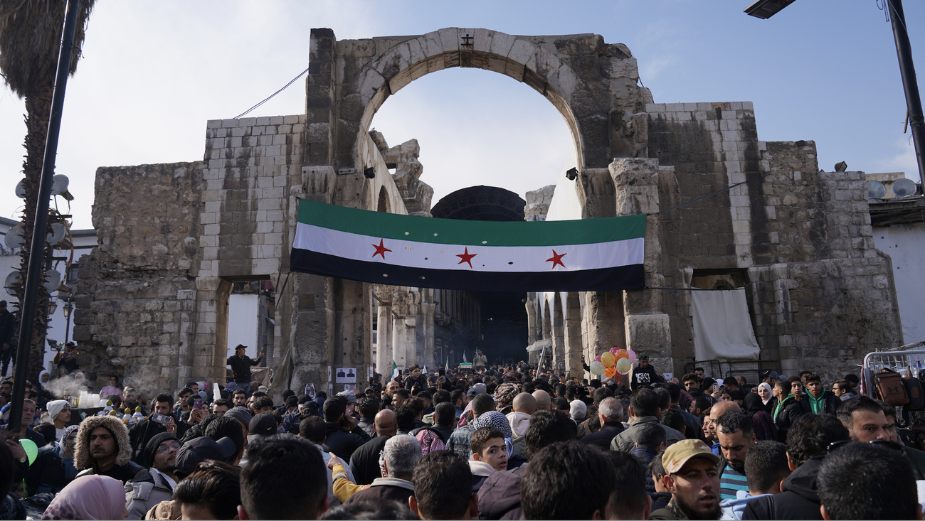 A huge crowd of people gathering outside, in front of a huge Syrian flag hanging from a gate. 