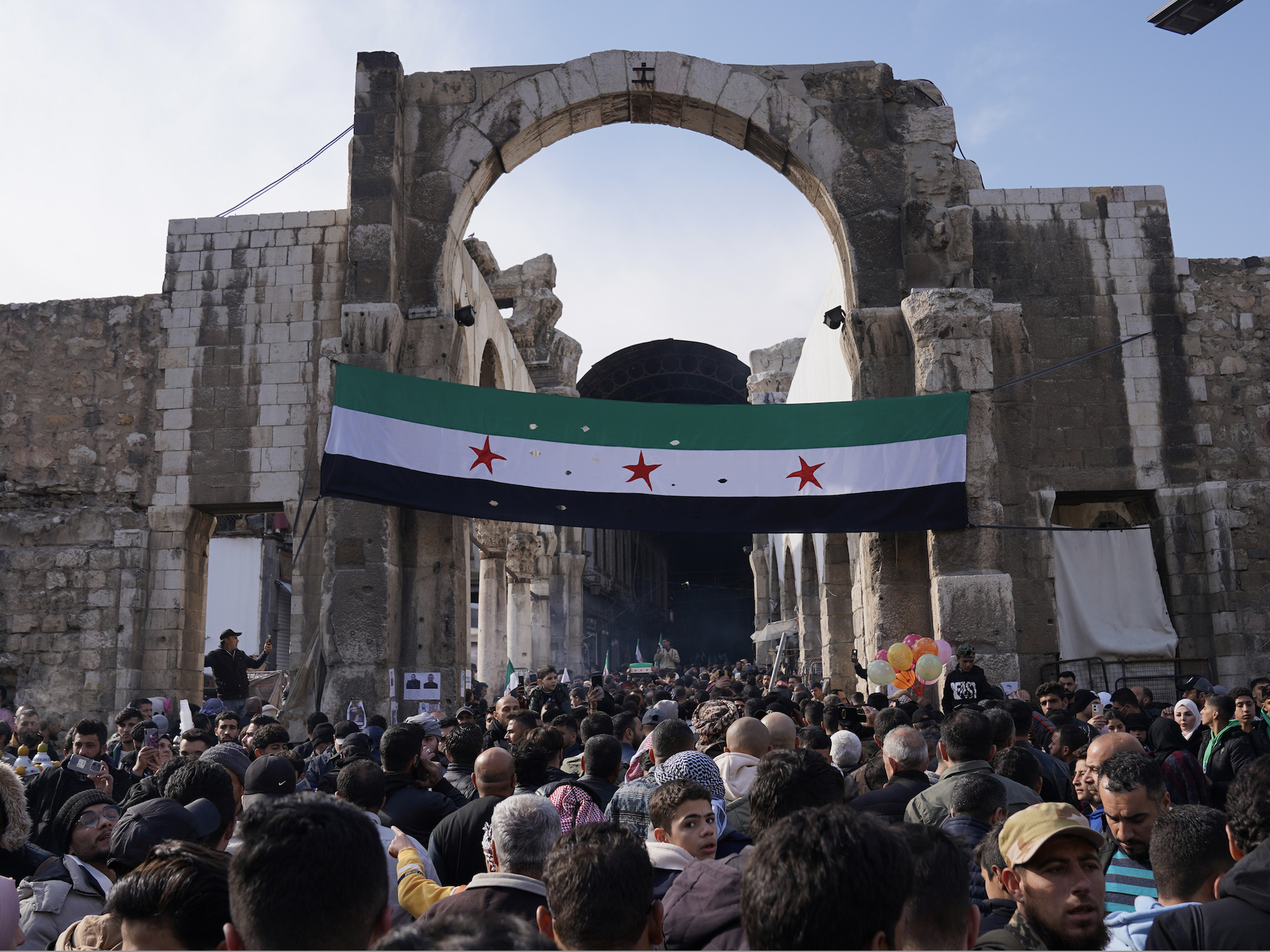 A huge crowd of people gathering outside, in front of a huge Syrian flag hanging from a gate. 